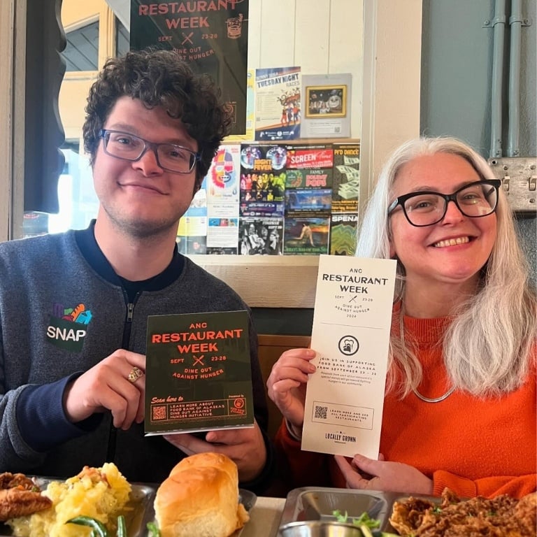 Two people sitting in a booth at Spenard Roadhouse, holding up Anchorage Restaurant Week signs and menus.