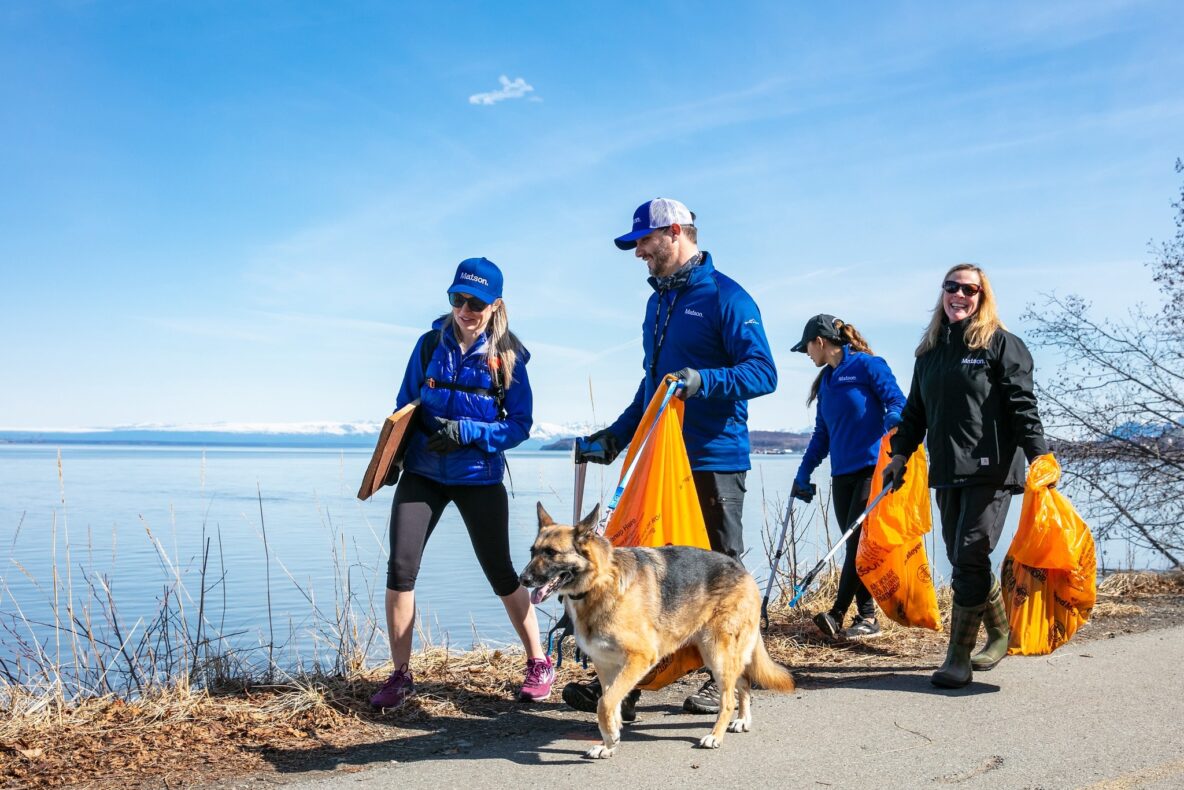 Four Matson employees wearing blue jackets and baseball hats walking along the coastal trail in early spring with trash pick-up bags and a dog. Turnagain Arm in the background and a bare trees in the foreground.